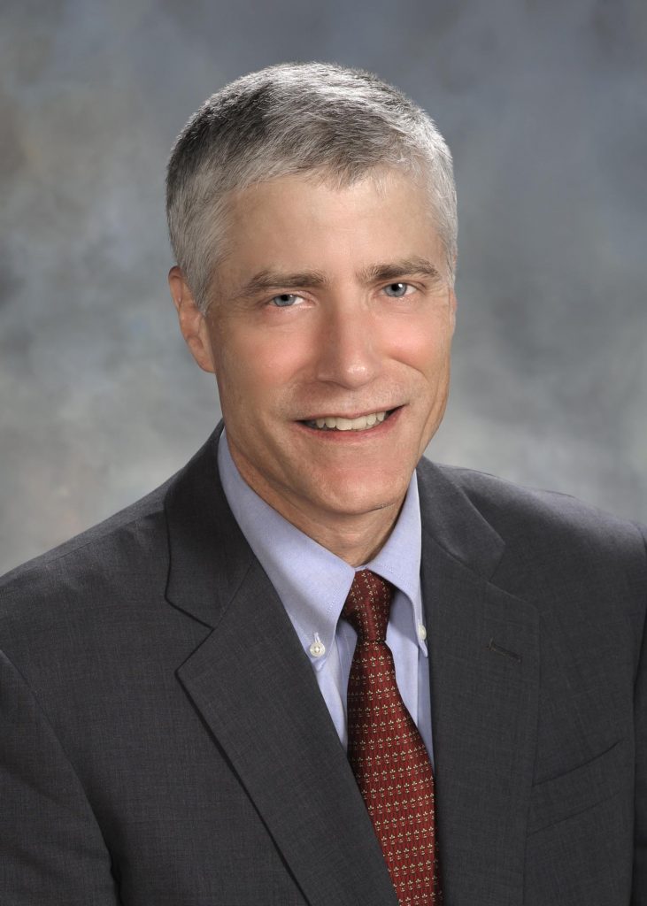 Professional photo of Dr. Netland.  He is wearing a grey suit coat, light blue dress shirt and a dark red tie. 
