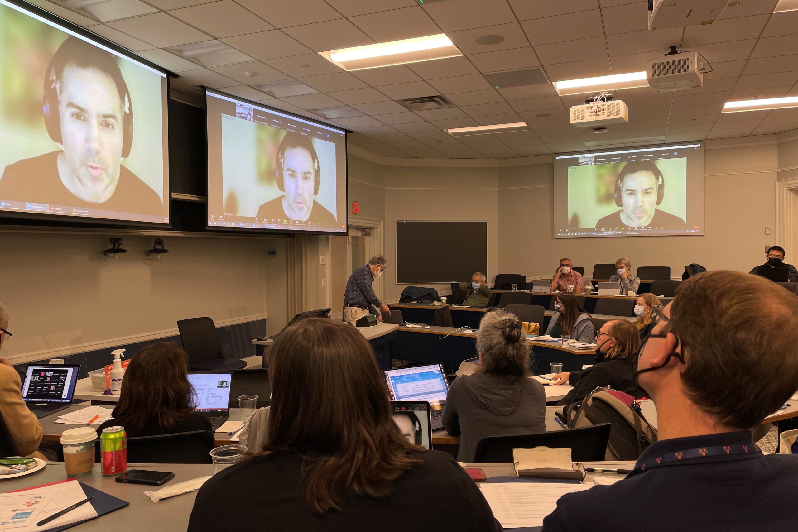 Neil Lagali's face is shown on three different screens at the front of conference room.  There are many in person attendees paying him rapt attention. 
