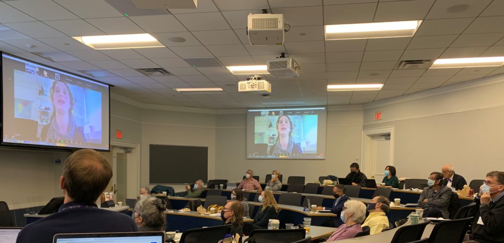 Nikki Hall is shown on two screens at the front and side of the conference room.  In person attendees are watching and listening to her. 