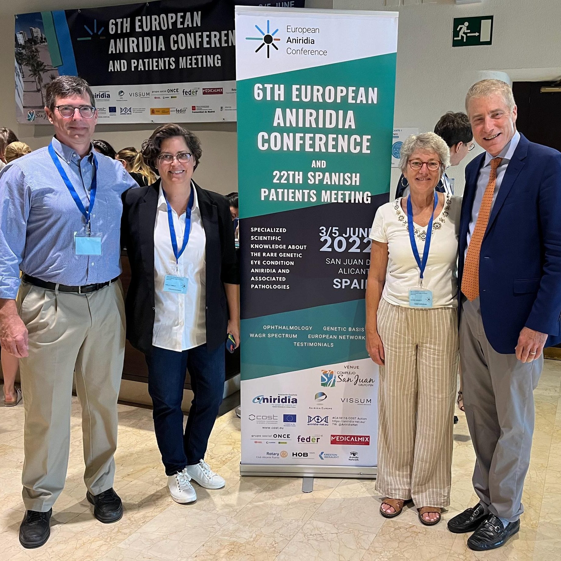 Picture is of 2 people standing next to each other, then a sign, then 2 more people.  From left is Jim Lauderdale, Kelly Trout, the 6th European Aniridia Conference sign, Shari Krantz, and Peter Netland.  