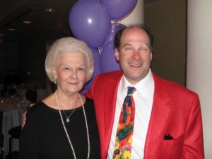 Candid photo of beautiful elderly woman on the left and middle aged man on the right.  Bobbi has silver hair and is wearing a black shirt with a long strand of pearls around her neck and a shorter pearl necklace.  Michael is wearing a read suit with a colorful tie and a white shirt.  Michael has his arm around his mom.  